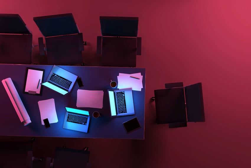 Desktop App Three active laptop computers on a desk inside an office space at night, with glowing blue light from monitors illuminating the table. Aerial view, as seen from above. Purple light, in contrast with blue displays, illuminates the dark background. Teamwork and deadlines. Tablets, smartphones, coffee mugs, paper documents and projects are also visible on the desk, surrounded by office chairs. Dark purple and red hues at midnight hour. Copy space on dark areas. Digitally generated image.