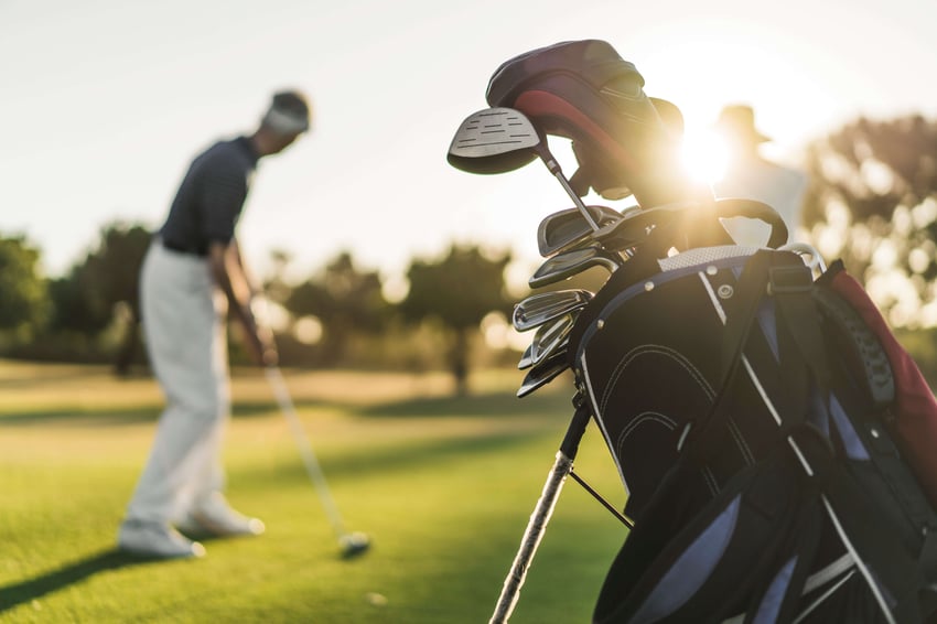 A photo of senior man and woman at golf course. Focus is on golf bag. They are playing on sunny day.